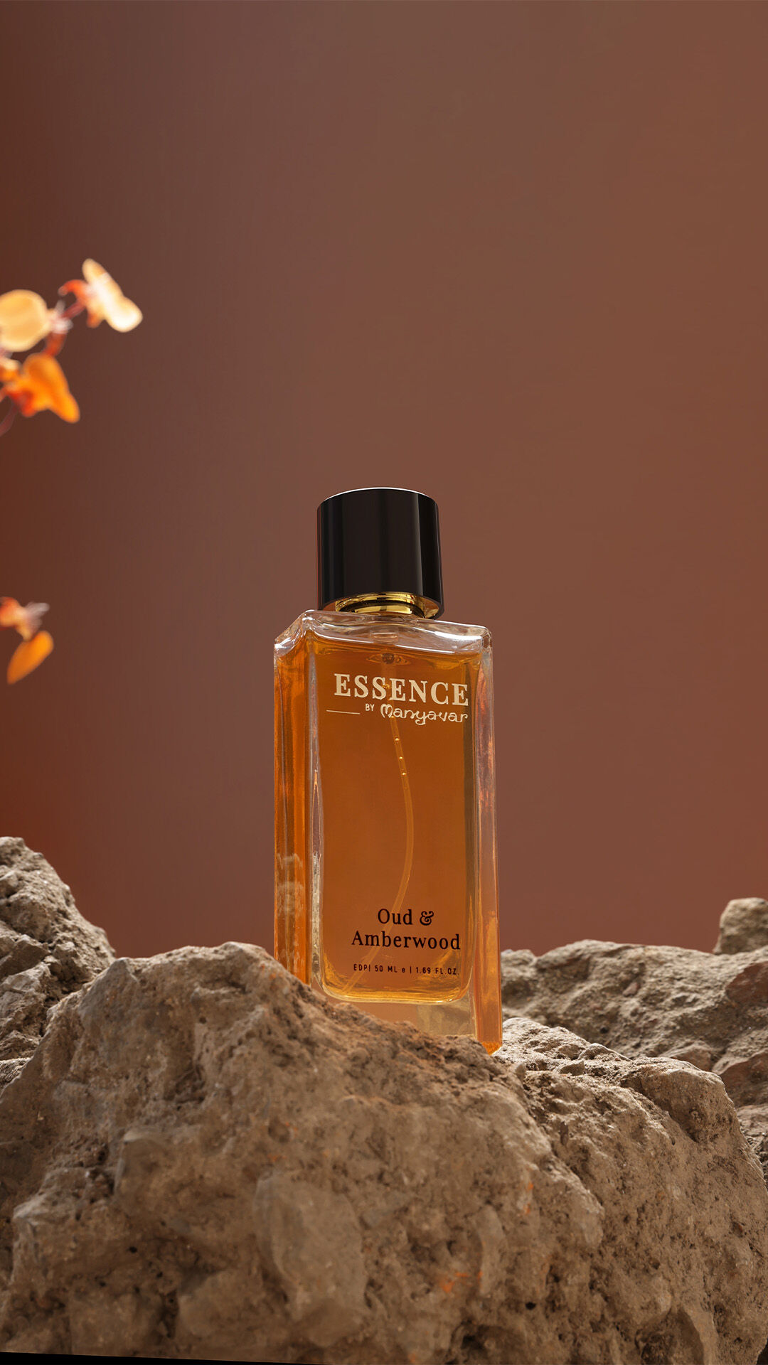 Essence by Manyavar Oud & Amberwood perfume bottle placed on rugged rocks with a warm brown backdrop and delicate orange floral accents